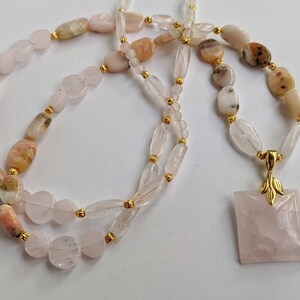 May include: A necklace featuring a variety of polished gemstones in shades of pink, peach, and cream, strung together with gold-colored beads. The necklace has a square rose quartz pendant with a gold leaf design.