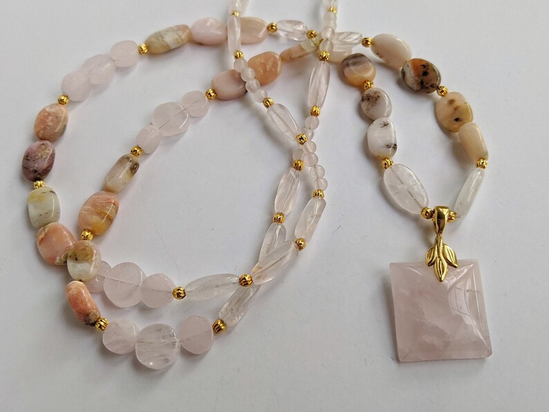 May include: A necklace featuring a variety of polished gemstones in shades of pink, peach, and cream, strung together with gold-colored beads. The necklace has a square rose quartz pendant with a gold leaf design.