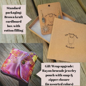 May include: A pair of earrings with a silver-colored floral design and gray stone beads, displayed in a brown cardboard box with cotton filling. The box has the logo "The Snarky Owl Workshop". A pink and gold brocade jewelry pouch is also shown.