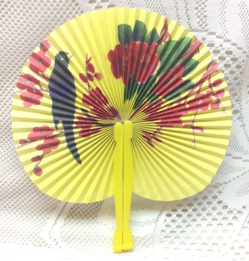 May include: A yellow folding fan with a black bird and red flowers painted on it. The fan is closed and has a yellow handle.