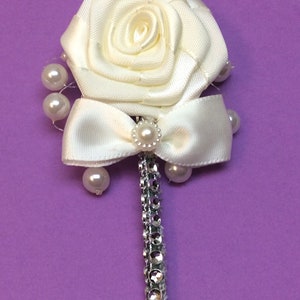 May include: A white satin rose with a white satin bow and pearl accents. The rose is attached to a silver rhinestone stem.