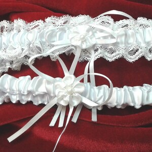 May include: Two white satin garters with lace trim and a white satin flower accent. The garters are on a red velvet background.