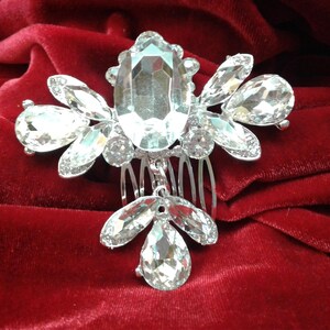 May include: A silver hair comb with a large clear crystal in the center and smaller crystals surrounding it. The comb is on a red velvet background.