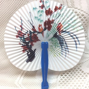 May include: A white folding fan with a floral design and blue handle. The fan features a painted design of a tree with red flowers and birds.
