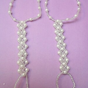 May include: A pair of white beaded foot jewelry with a delicate design. Each piece has a circular ankle bracelet and a long beaded strand that ends in a loop.