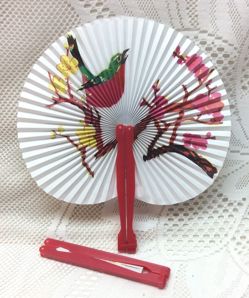 May include: A white folding fan with a red, green, and yellow bird on a branch with pink flowers. The fan has a red handle.