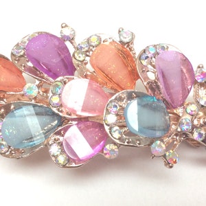 May include: A gold hair clip with a decorative design featuring multiple teardrop-shaped gems in shades of pink, blue, and orange. The gems are surrounded by smaller clear rhinestones.