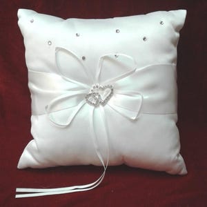 May include: White satin pillow with a white satin bow and two interlocking rhinestone hearts. The pillow has a long white satin ribbon attached.