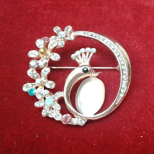 May include: A gold-tone brooch featuring a peacock design with a white iridescent stone and a circle of clear rhinestones. The peacock is surrounded by a ring of flowers with colorful rhinestones.