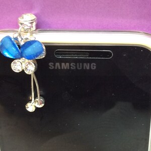 May include: A blue butterfly charm with rhinestones attached to a silver phone charm. The charm is attached to a Samsung phone.