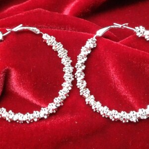 May include: A pair of silver hoop earrings with a textured, twisted design. The earrings are on a red velvet background.