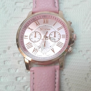May include: A rose gold analog wristwatch with a white face and Roman numeral hour markers. The watch has a pink leather band and three smaller dials on the face. The watch face says "Geneva" and "Quartz".