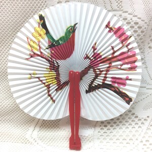 May include: A white folding fan with a red, green, and black bird design and pink flowers. The fan has a red plastic handle.