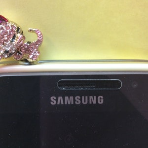 May include: A pink and gold jeweled cat charm sits on top of a white Samsung phone case.