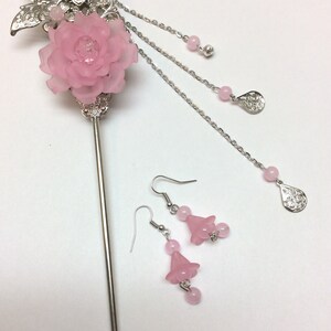 May include: A silver hair stick with a pink flower and butterfly design. The stick has three dangling chains with pink beads and silver charms. A pair of pink flower earrings with pink beads hang below.
