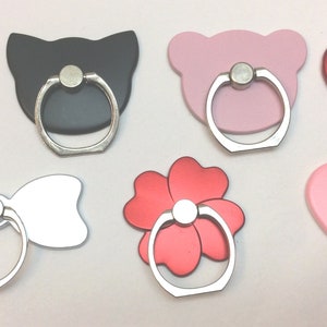 May include: Assortment of phone ring holders in various shapes and colors, including a flower, a bow, a heart, and a cat. The holders are made of metal and have a silver ring.