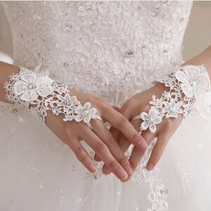 May include: A pair of white lace bridal gloves with delicate floral designs and sparkling rhinestones. The gloves are fingerless and have a wrist strap.