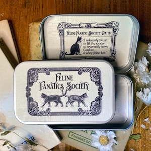 Könnte beinhalten: Zwei rechteckige silberne Metalldosen mit Deckeln. Der obere Deckel zeigt den Text Feline Fanatics Society mit zwei Katzen. Der geöffnete Deckel zeigt den Text Feline Fanatics Society Oath und eine Katzenillustration. Die Dosen sind metallisch.