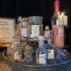 May include: A collection of clear glass apothecary bottles with labels reading Adder's Fork, Eye of Newt, Blind Worm's Sting, Wing of Bat, and Toe of Frog, arranged on a metal tray with a framed Midnight Margaritas sign and a bottle of Diablo de Flores.