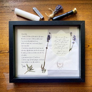 May include: A framed artwork with handwritten letters, dried lavender sprigs, and an envelope. The black frame surrounds the artwork, which includes a white candle, a wax stamp, and a bundle of lavender. The text is in black ink on cream-coloured paper.