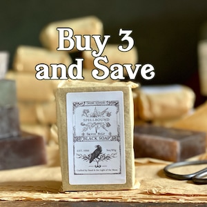 May include: A bar of Spellbound Black Soap wrapped in brown paper. The soap is labeled with a black raven and the text "Savon Noir" and "Black Soap". The text "Buy 3 and Save" is written in white letters on a green background.