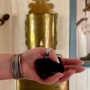 Op de afbeelding: Een zwart, rechthoekig object wordt vastgehouden in een hand versierd met zilveren ringen en meerdere gouden en zilveren armbanden. Een vintage messing wandcontainer staat op de achtergrond.