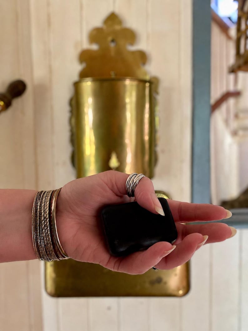 Op de afbeelding: Een zwart, rechthoekig object wordt vastgehouden in een hand versierd met zilveren ringen en meerdere gouden en zilveren armbanden. Een vintage messing wandcontainer staat op de achtergrond.