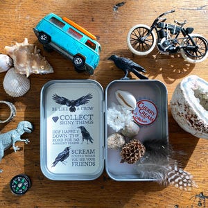 Puede incluir: Una colecci&oacute;n de objetos peque&ntilde;os sobre una superficie de madera, que incluye una furgoneta de juguete azul, una motocicleta de juguete, conchas marinas, una geoda, una figura de dinosaurio, una br&uacute;jula y una lata con el texto "BE LIKE A CROW COLLECT SHINY THINGS".