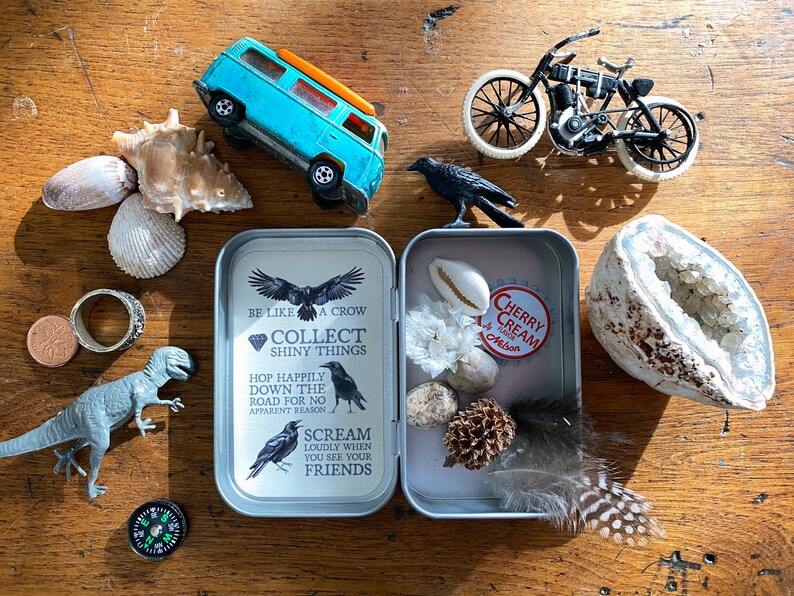 Puede incluir: Una colecci&oacute;n de objetos peque&ntilde;os sobre una superficie de madera, que incluye una furgoneta de juguete azul, una motocicleta de juguete, conchas marinas, una geoda, una figura de dinosaurio, una br&uacute;jula y una lata con el texto "BE LIKE A CROW COLLECT SHINY THINGS".