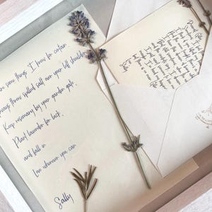 May include: A framed artwork featuring handwritten letters and dried lavender sprigs. The letters, in elegant cursive, include phrases like "Love whenever you can" and "Plant lavender for luck." The artwork is presented in a white wooden frame.