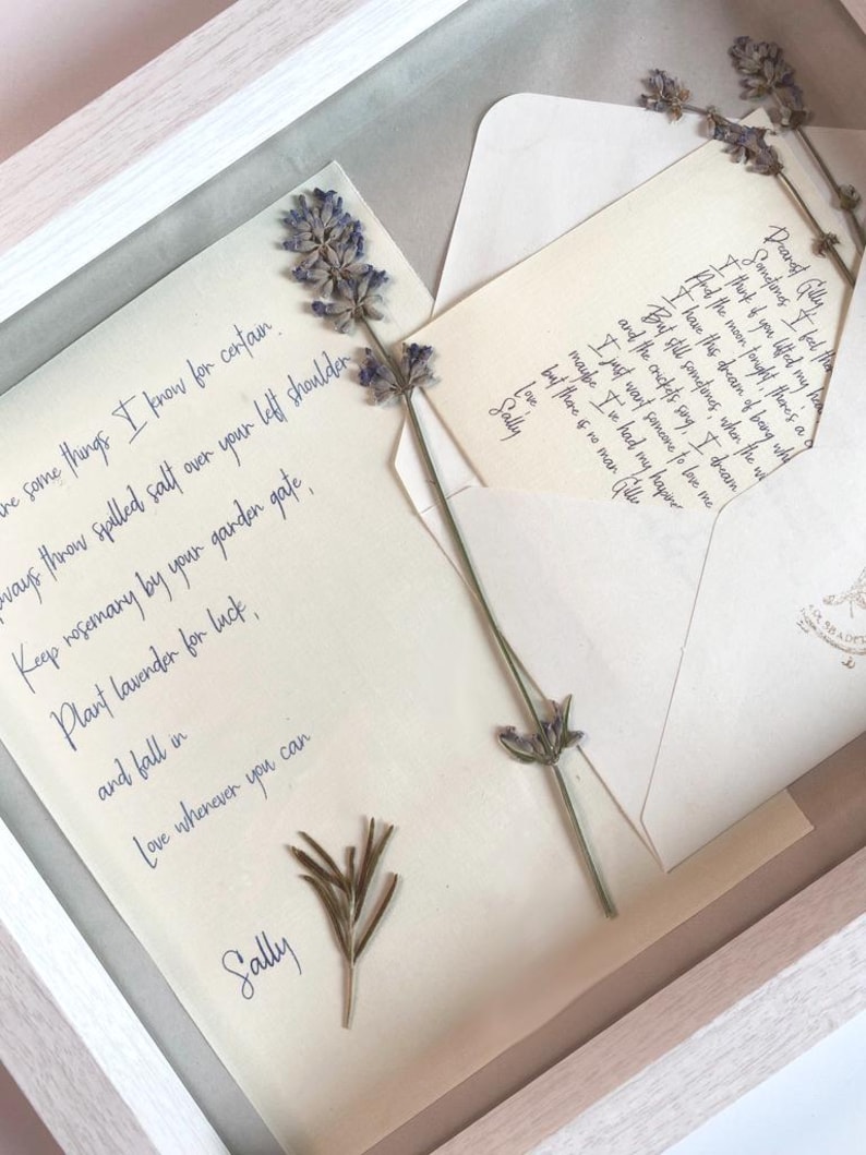 May include: A framed artwork featuring handwritten letters and dried lavender sprigs. The letters, in elegant cursive, include phrases like "Love whenever you can" and "Plant lavender for luck." The artwork is presented in a white wooden frame.