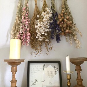 May include: A rustic display featuring dried flower bouquets in shades of beige, pink, white, and purple, hanging from a wooden rack. A framed botanical print and candles in wooden and brass holders complete the decor.