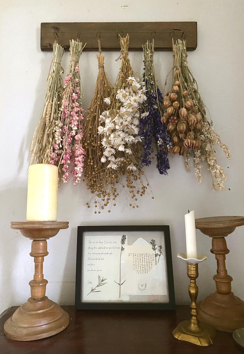 May include: A rustic display featuring dried flower bouquets in shades of beige, pink, white, and purple, hanging from a wooden rack. A framed botanical print and candles in wooden and brass holders complete the decor.