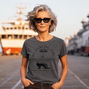 May include: A charcoal gray short-sleeved t-shirt featuring a black graphic print. The design includes an umbrella, a vintage shoe, and the text: My Darling Girl, When are you going to understand that being normal is not necessarily a virtue? It rather denotes a lack of courage. Aunt Frances - Practical Magic.