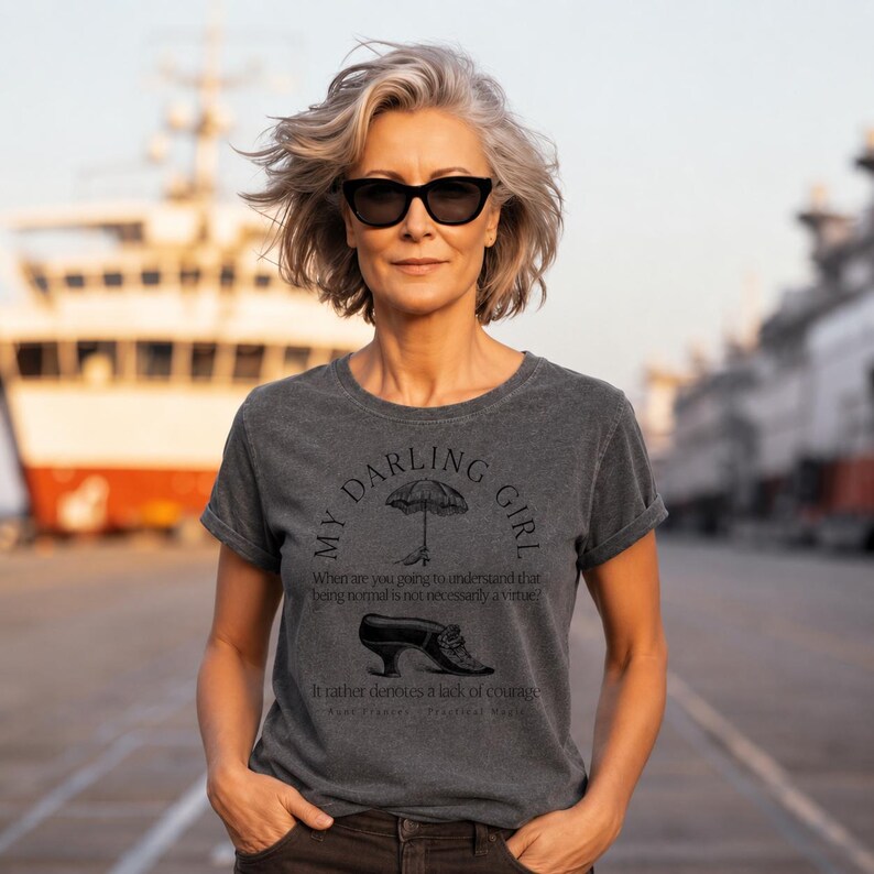 May include: A charcoal gray short-sleeved t-shirt featuring a black graphic print. The design includes an umbrella, a vintage shoe, and the text: My Darling Girl, When are you going to understand that being normal is not necessarily a virtue? It rather denotes a lack of courage. Aunt Frances - Practical Magic.