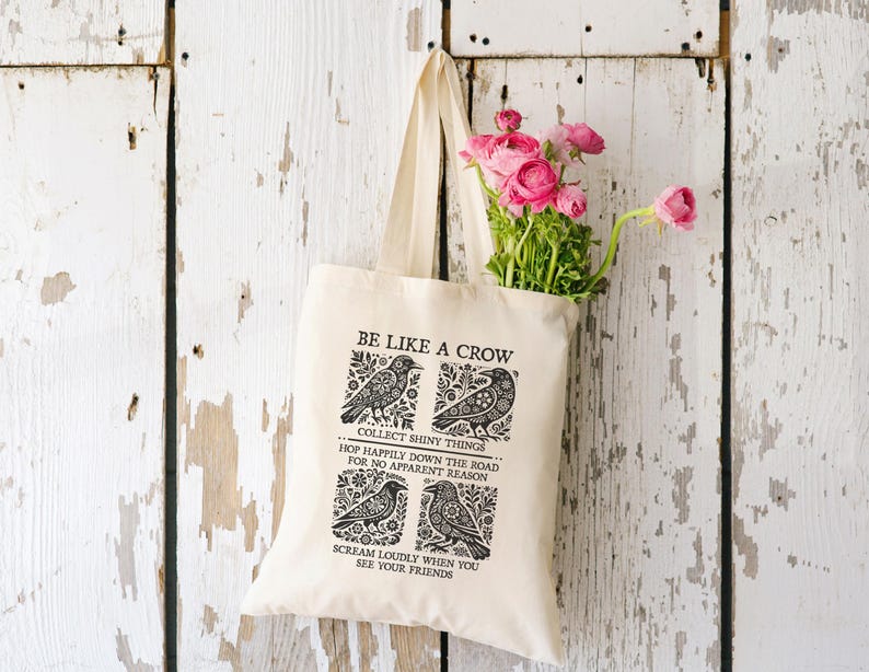 May include: A cream-colored tote bag with the text "BE LIKE A CROW" and crow illustrations. The bag contains pink roses and is hanging against a weathered white wooden background.
