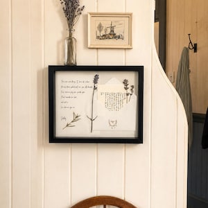 May include: A framed display featuring a handwritten letter, dried lavender, and an envelope with script. A small framed print of a windmill and a vase of lavender sprigs are also visible. The items are displayed on a white paneled wall.