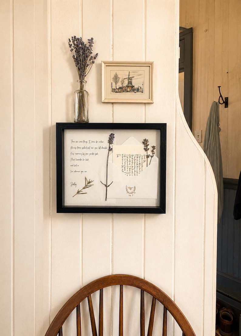 May include: A framed display featuring a handwritten letter, dried lavender, and an envelope with script. A small framed print of a windmill and a vase of lavender sprigs are also visible. The items are displayed on a white paneled wall.
