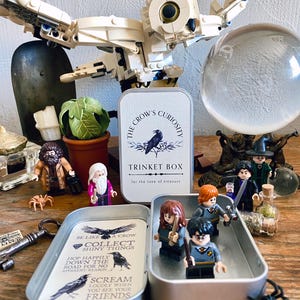 Puede incluir: Una caja de baratijas de metal con el texto "The Crow's Curiosity" y una ilustraci&oacute;n de cuervo. En el interior, se exhiben figuras en miniatura de personajes de una serie de fantas&iacute;a. Tambi&eacute;n hay un b&uacute;ho de Lego y otros objetos decorativos.