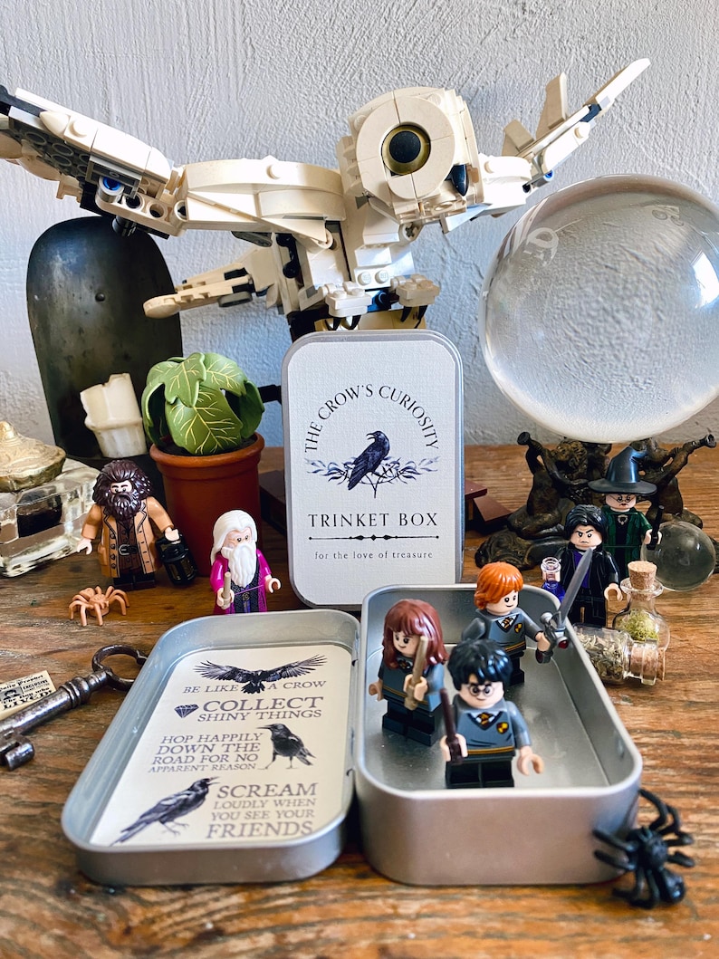Puede incluir: Una caja de baratijas de metal con el texto "The Crow's Curiosity" y una ilustraci&oacute;n de cuervo. En el interior, se exhiben figuras en miniatura de personajes de una serie de fantas&iacute;a. Tambi&eacute;n hay un b&uacute;ho de Lego y otros objetos decorativos.
