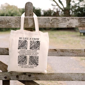 Puede incluir: Un bolso tote de color crema con el texto "BE LIKE A CROW" e ilustraciones de cuervos. La bolsa tambi&eacute;n presenta las frases "COLLECT SHINY THINGS", "HOP HAPPILY DOWN THE ROAD FOR NO APPARENT REASON" y "SCREAM LOUDLY WHEN YOU SEE YOUR FRIENDS".