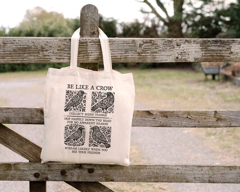 Puede incluir: Un bolso tote de color crema con el texto "BE LIKE A CROW" e ilustraciones de cuervos. La bolsa tambi&eacute;n presenta las frases "COLLECT SHINY THINGS", "HOP HAPPILY DOWN THE ROAD FOR NO APPARENT REASON" y "SCREAM LOUDLY WHEN YOU SEE YOUR FRIENDS".