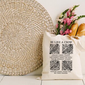 Puede incluir: Una bolsa de mano crema con el texto "BE LIKE A CROW" e ilustraciones de cuervos. Contiene flores rosas y dos baguettes. Un gran salvamanteles redondo tejido se encuentra en el fondo.