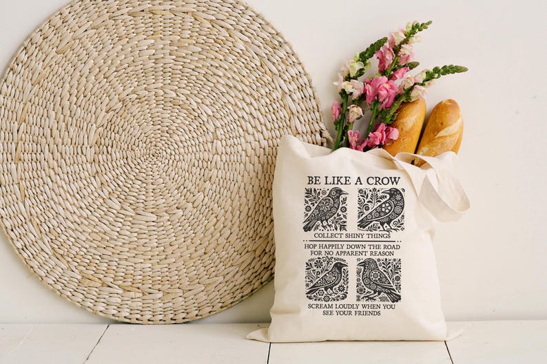 Puede incluir: Una bolsa de mano crema con el texto "BE LIKE A CROW" e ilustraciones de cuervos. Contiene flores rosas y dos baguettes. Un gran salvamanteles redondo tejido se encuentra en el fondo.