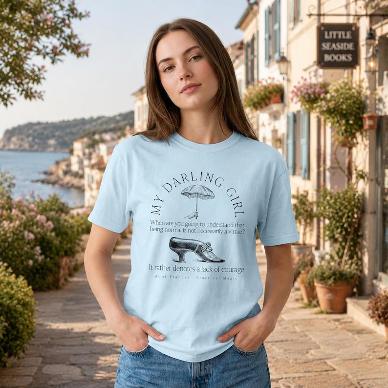 May include: A light blue t-shirt featuring a black graphic print of an umbrella and a vintage shoe. The text reads: MY DARLING GIRL, When are you going to understand that being normal is not necessarily a virtue? It rather denotes a lack of courage, Aunt Frances, Practical Magic.