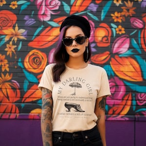 May include: A beige t-shirt featuring black text that reads MY DARLING GIRL, an illustration of an umbrella, and a shoe. Below, the text reads: When are you going to understand that being normal is not necessarily a virtue? It rather denotes a lack of courage.