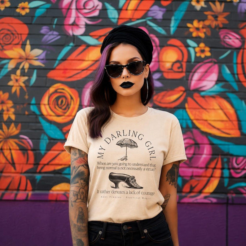 May include: A beige t-shirt featuring black text that reads MY DARLING GIRL, an illustration of an umbrella, and a shoe. Below, the text reads: When are you going to understand that being normal is not necessarily a virtue? It rather denotes a lack of courage.