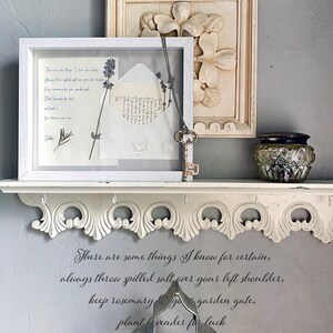May include: A white framed display with a pressed lavender sprig and an open envelope. A decorative shelf holds the frame, a carved floral relief, and a small glass jar. A metal key hangs from the frame. The wall features a quote in script.