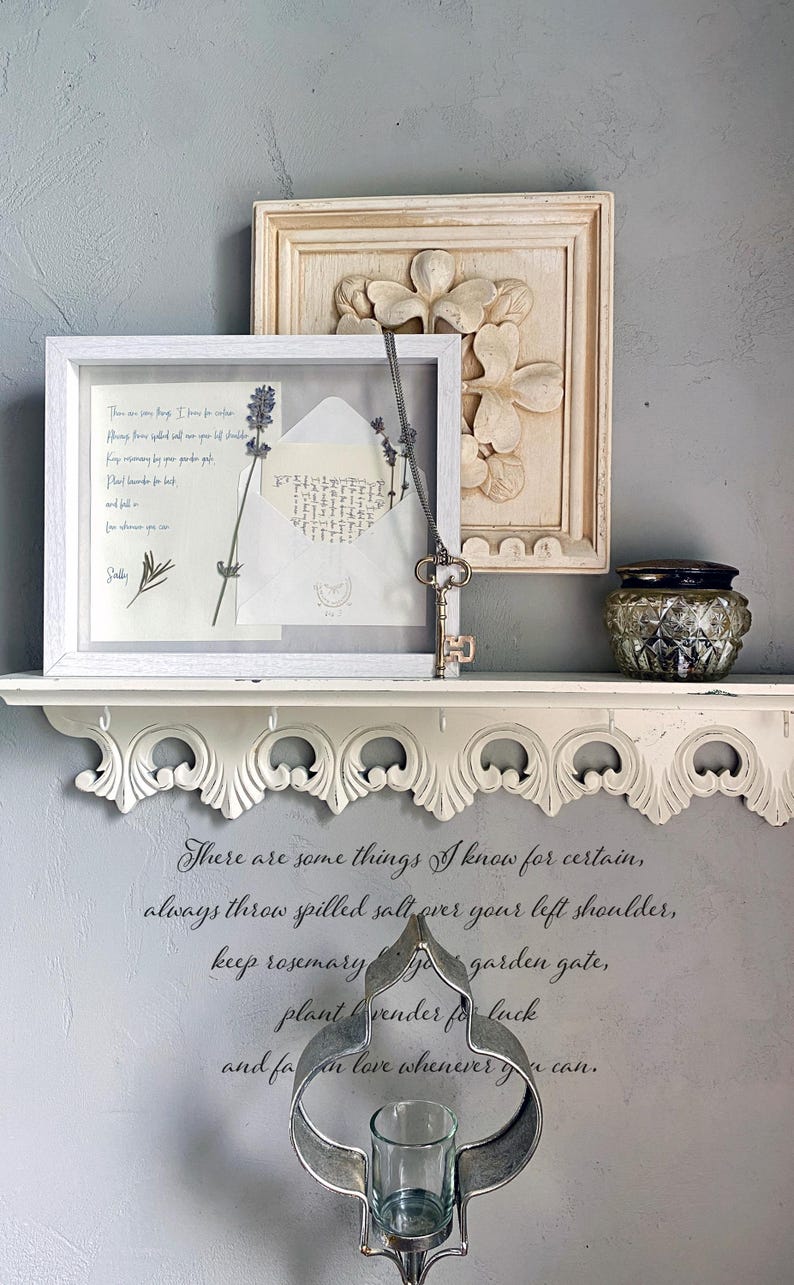 May include: A white framed display with a pressed lavender sprig and an open envelope. A decorative shelf holds the frame, a carved floral relief, and a small glass jar. A metal key hangs from the frame. The wall features a quote in script.