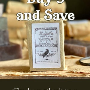 Op de afbeelding: Een stuk zwarte zeep verpakt in bruin papier met een etiket met de tekst "Spellbound Black Soap". Het etiket toont een illustratie van een raaf en de tekst "Buy 3 and Save". De zeep weegt 85g.